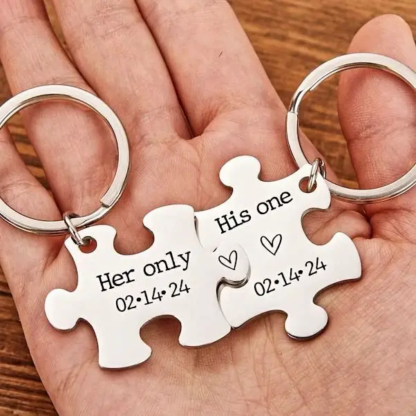 Two interlocking puzzle piece keychains with engravings, one saying "Her only" with a date, and the other "His one" with a date, held in a hand.