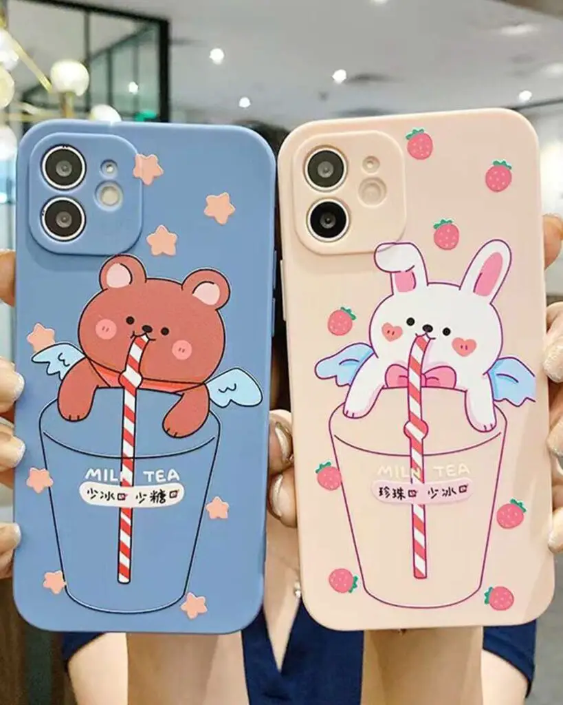 Two hands holding smartphones with cases depicting a bear and a bunny enjoying milk tea, embellished with stars and strawberries.