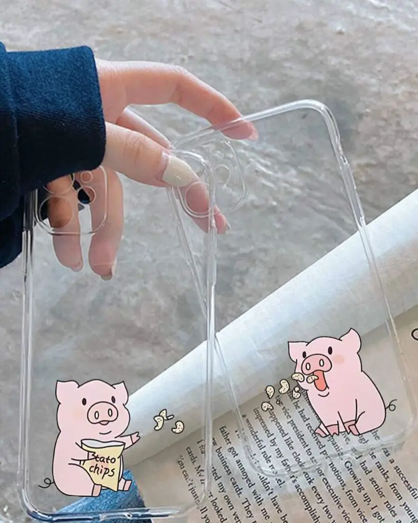 A hand holding a transparent smartphone case over a book, with a cartoon pig enjoying "Bato chips" illustrated on the bottom corner of the case.