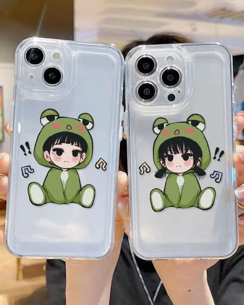 Two iPhone cases with cute anime-style characters in green frog costumes, complete with musical notes, showcased against a blurry indoor background.