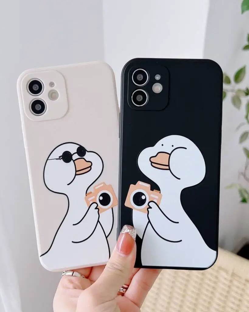 A pair of iPhone cases showing a cartoon duck taking a selfie, one on a white background and the other on black, held in a person's hands.