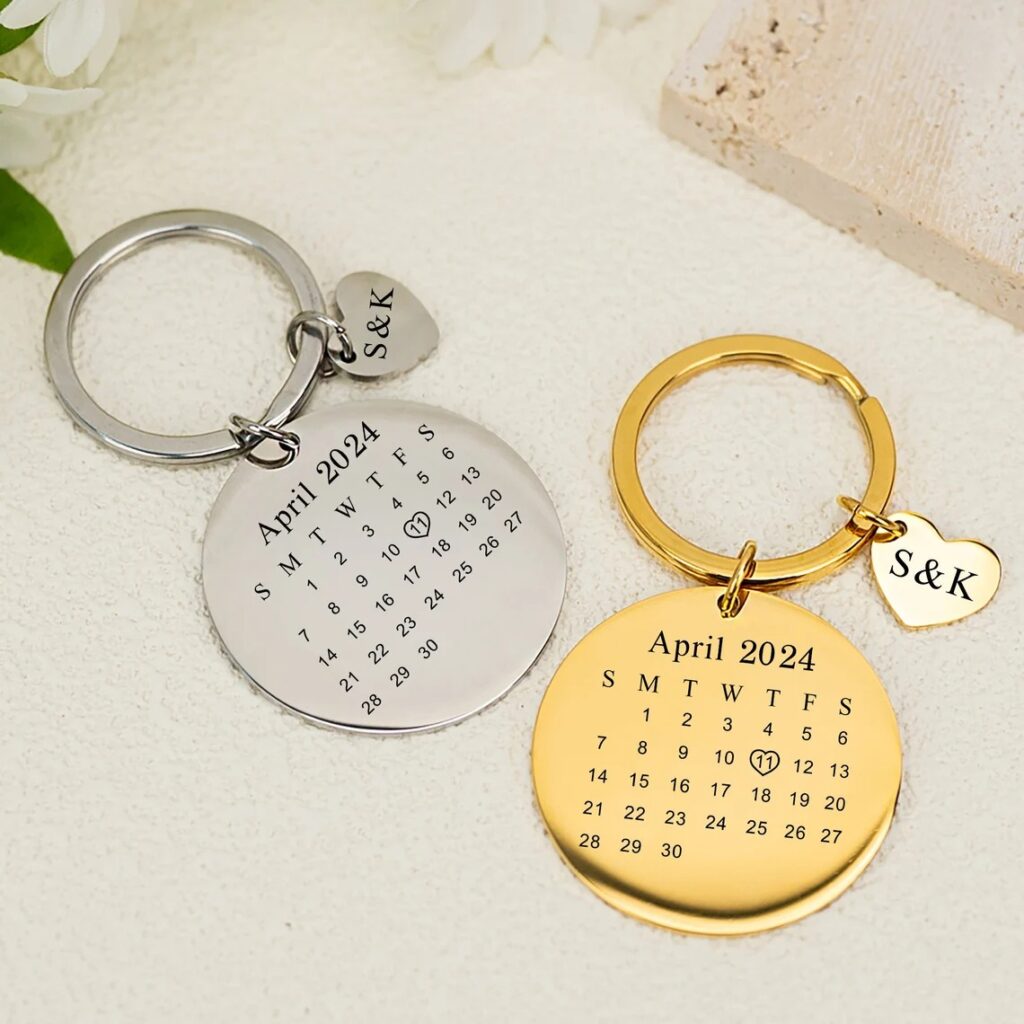 Silver and gold round keychains with the calendar of April 2024 and initials on heart charms, displayed on a floral background.