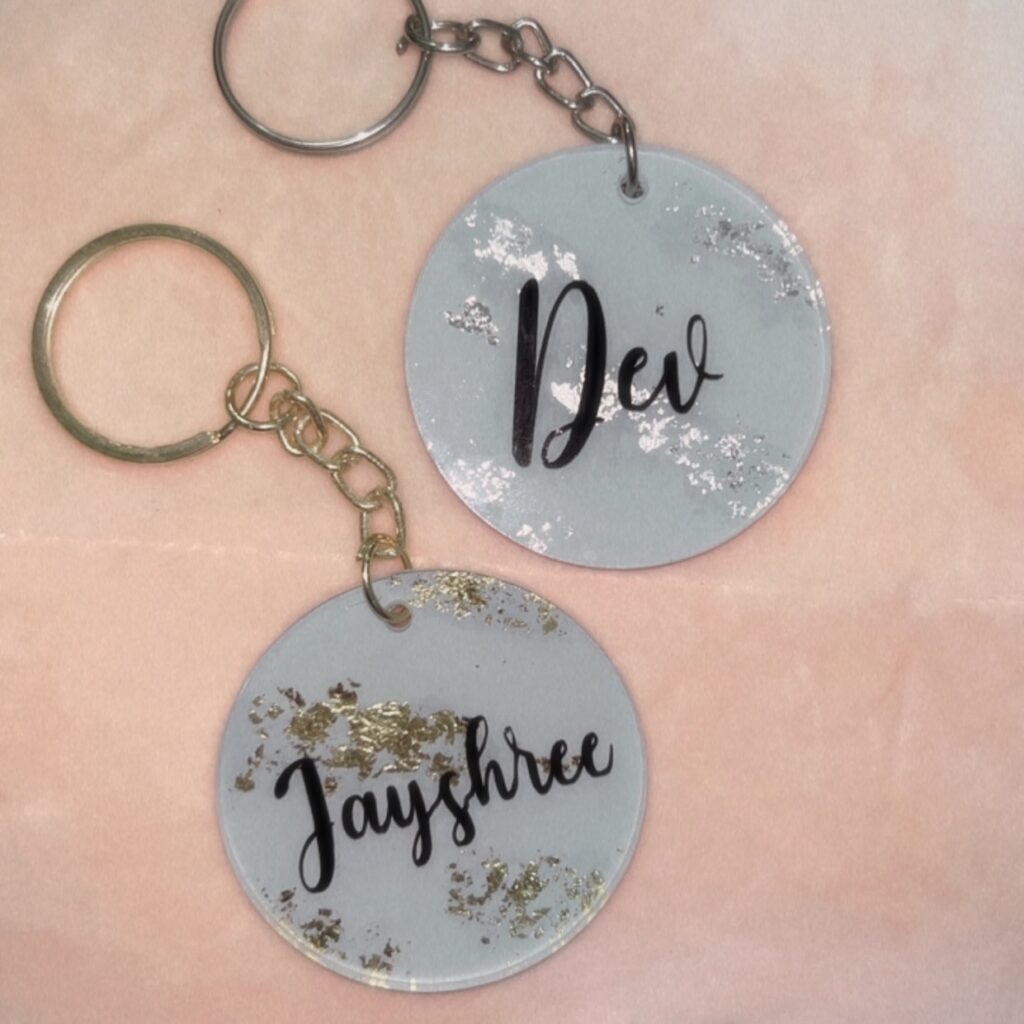 Two circular keychains with names "Dev" and "Jayshree" in black over a blue background with gold and silver flakes.