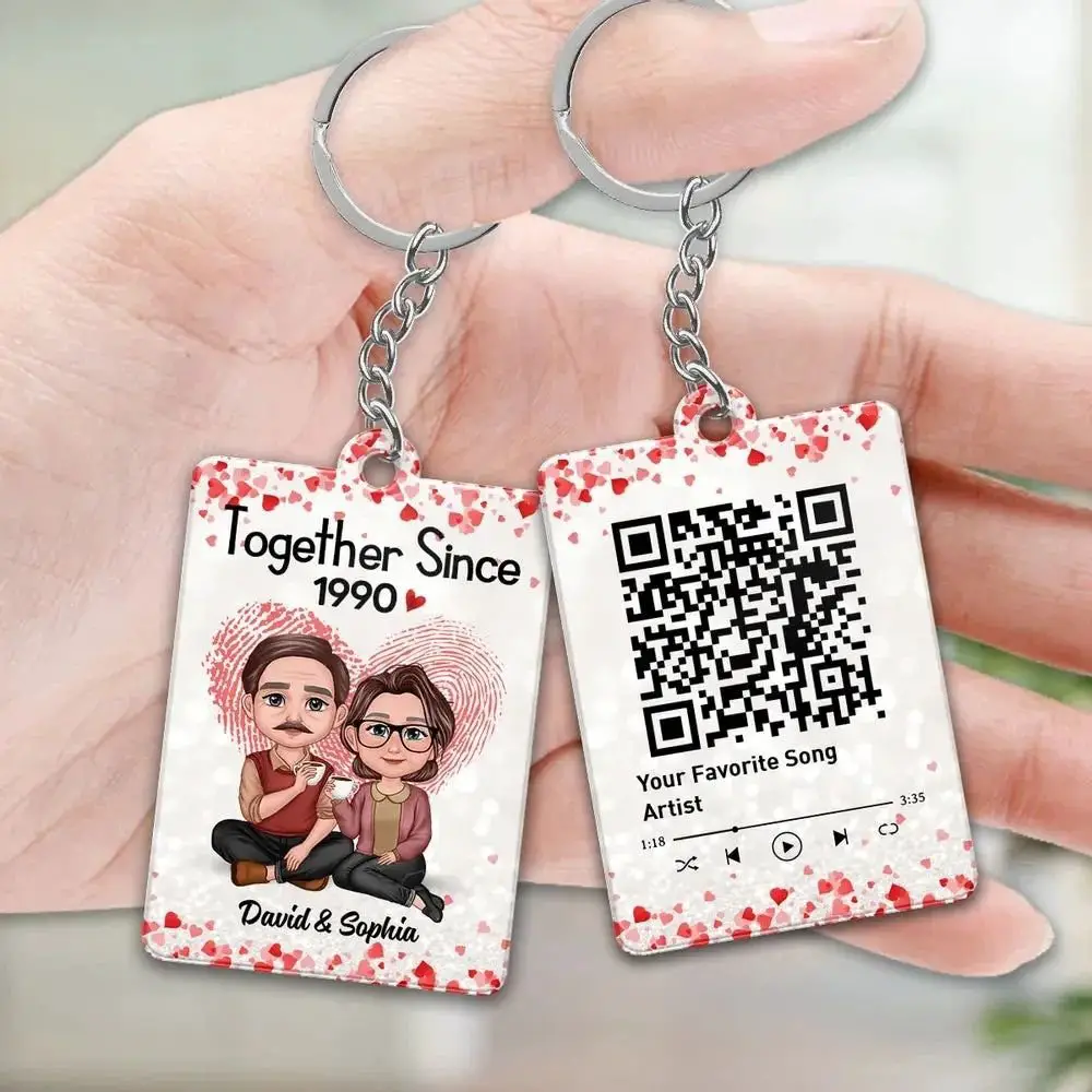Two keychains held in hand, one illustrated with a couple and the phrase "Together Since 1990," and the other displaying a QR code for a song.