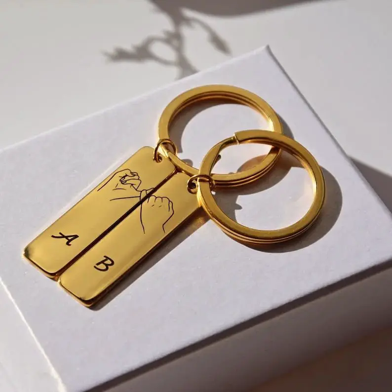 A golden keychain with a hand gesture drawing forming a heart and initials, resting on a white surface.