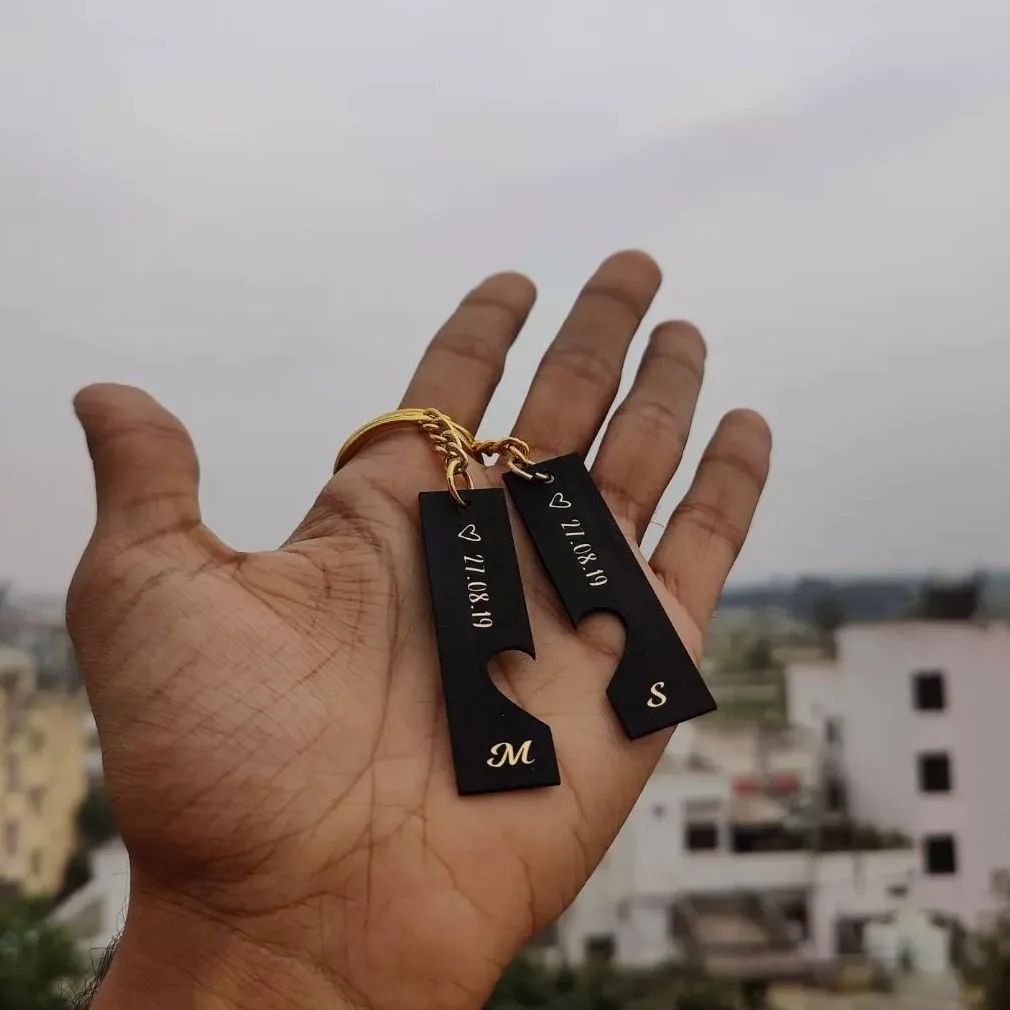Black rectangular keychains with engraved dates and initials, held in hand with a cityscape background.
