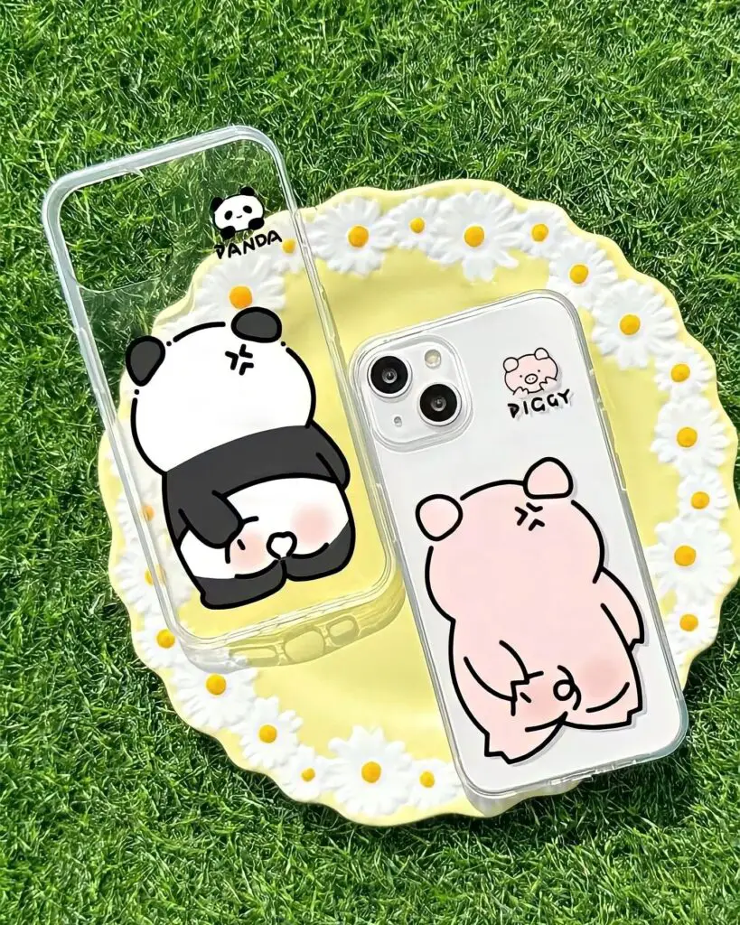 Transparent smartphone cases held over grass, featuring cartoon panda and pig characters, surrounded by small white flowers.