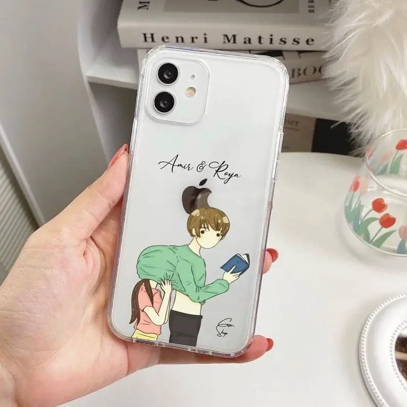 A transparent iPhone case with a cartoon illustration of a young couple, labeled with the names 'Anis & Roya', against a backdrop of books and decor.
