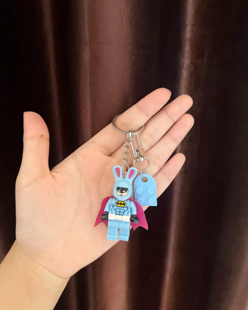 A mini figure keychain of a superhero with a bunny mask and pink cape, held in a hand against a dark curtain background.