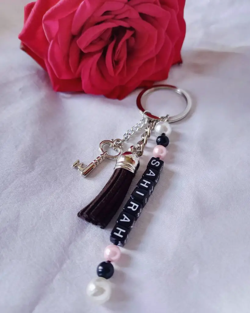 A custom keychain with silver charms, black and pink beads spelling 'SHIRA', and a red rose in the background.