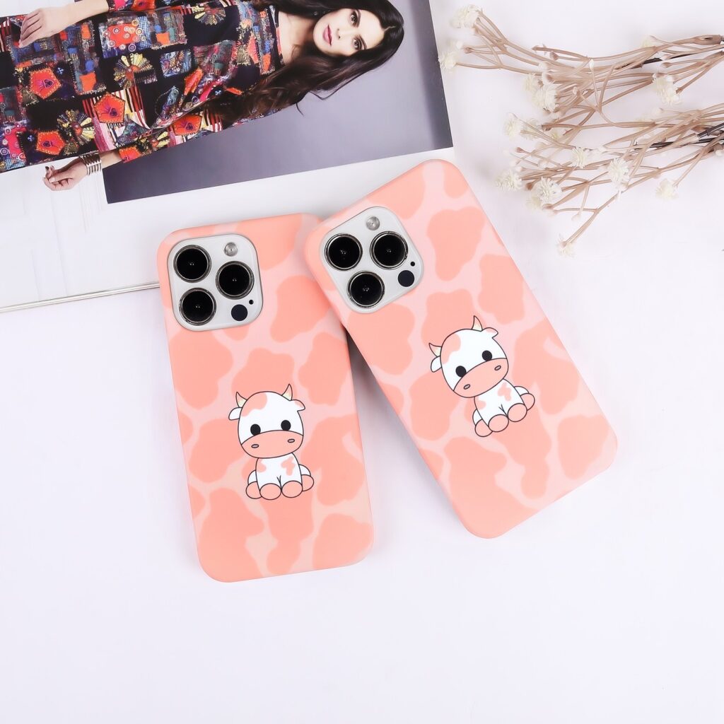 Two iPhone cases with cartoon cow designs on a pink camouflage background, displayed alongside a fashion magazine and decorative flowers.